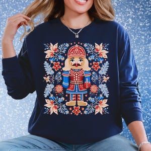 May include: Navy blue sweatshirt featuring a colorful nutcracker design. The graphic includes a detailed nutcracker figure in red, blue, and gold, surrounded by floral and leaf patterns. The long-sleeved top is a comfortable, casual style.
