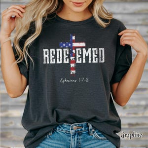 May include: Dark gray t-shirt featuring a cross design in the colors of the American flag, with the word "REDEEMED" in white block letters. The text "Ephesians 1:7-8" is below the cross. The shirt is made of a soft material.