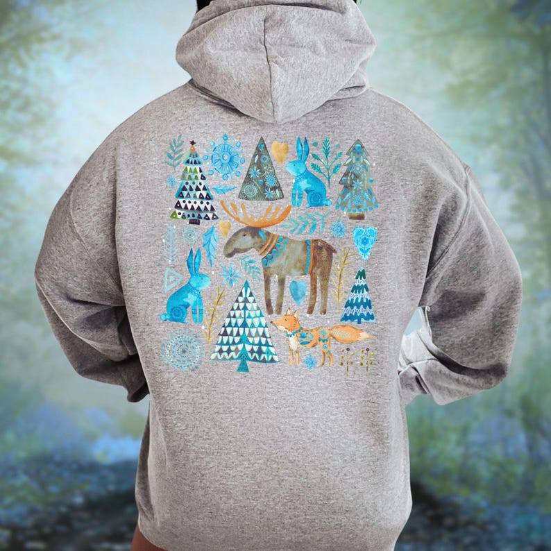 May include: A gray hoodie featuring a whimsical winter design. The back of the hoodie showcases a watercolor illustration of a moose, rabbits, Christmas trees, and a fox in shades of blue, brown, and orange. The design is set against a backdrop of a winter forest.