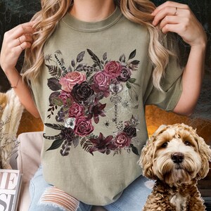 May include: A green t-shirt with a floral design featuring dark pink, burgundy, and black roses, leaves, and feathers. A silver key and spider web are in the center of the design.