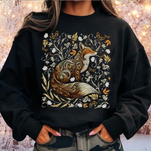 May include: Black sweatshirt featuring a detailed illustration of a fox with intricate golden patterns. The fox is surrounded by floral and butterfly designs in gold and white. The sweatshirt has long sleeves and a crew neck.