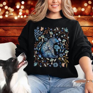 May include: A black sweatshirt with a blue watercolor illustration of a bear sitting in a forest scene. The bear is surrounded by flowers, mushrooms, and butterflies.