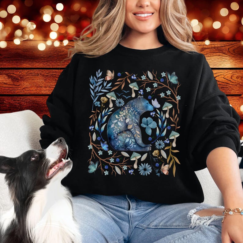 May include: A black sweatshirt with a blue watercolor illustration of a bear sitting in a forest scene. The bear is surrounded by flowers, mushrooms, and butterflies.