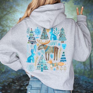 May include: A light gray hoodie featuring a winter-themed design on the back. The design includes blue and white illustrations of a moose, rabbits, Christmas trees, and a fox. The hoodie has a hood and long sleeves.