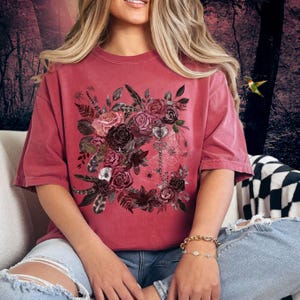 May include: A red oversized t-shirt with a floral design featuring dark red, pink, and white roses, leaves, and feathers. A key and spiderweb are also included in the design.