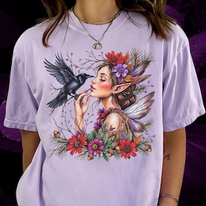 May include: A light purple t-shirt featuring a detailed illustration of a fairy with a raven. The fairy has long ears, wings, and is adorned with flowers. The raven is perched on a branch near the fairy's face, surrounded by red and purple flowers.