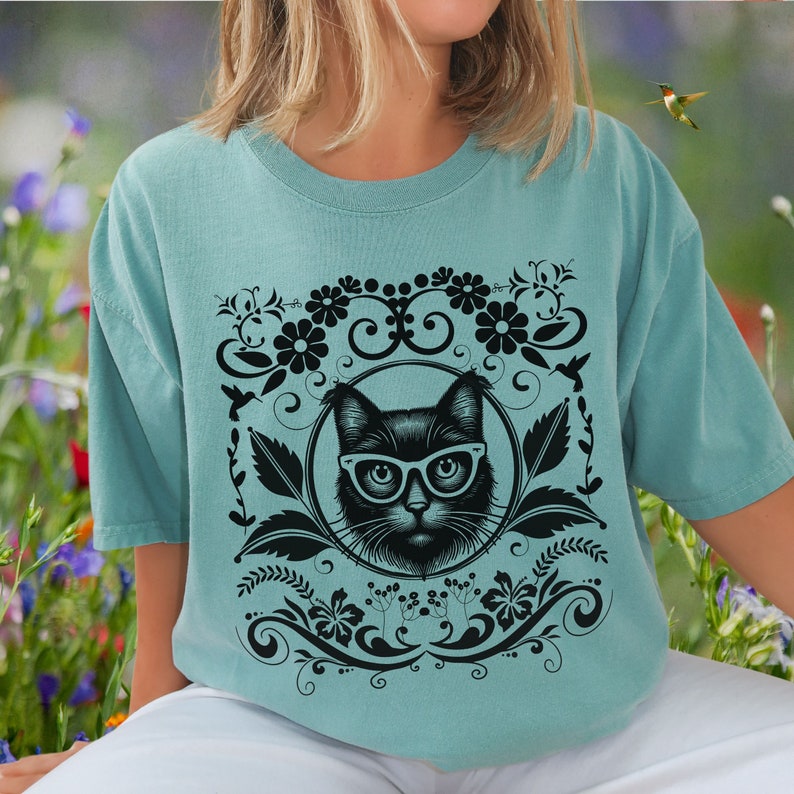 May include: A teal blue t-shirt with a black cat wearing glasses and a floral design. The cat is surrounded by a circular frame with a floral design.