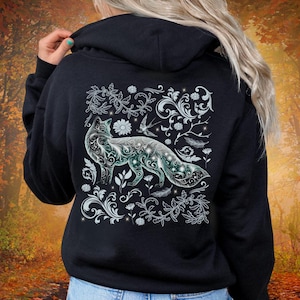 May include: Black hoodie featuring a detailed fox design on the back. The fox is depicted with intricate floral and leaf patterns in white and teal, surrounded by additional floral elements and feathers. The hoodie has a hood and a relaxed fit.