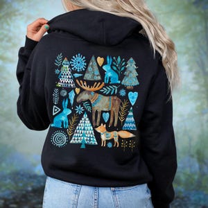 May include: Black hoodie featuring a whimsical winter design. The back of the hoodie displays a pattern of blue rabbits, trees, snowflakes, and a moose. The design incorporates teal, gold, and white accents, creating a festive and nature-inspired aesthetic.