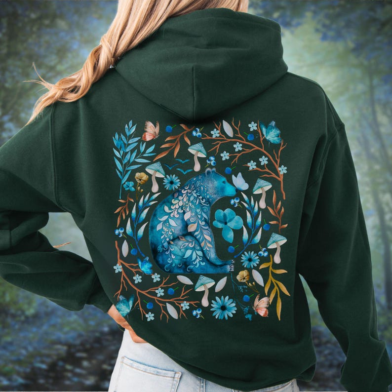 May include: Dark green hoodie featuring a blue bear design surrounded by flowers, mushrooms, and butterflies. The bear is intricately detailed with floral patterns. The design is centered on the back of the hoodie, creating a nature-inspired aesthetic.