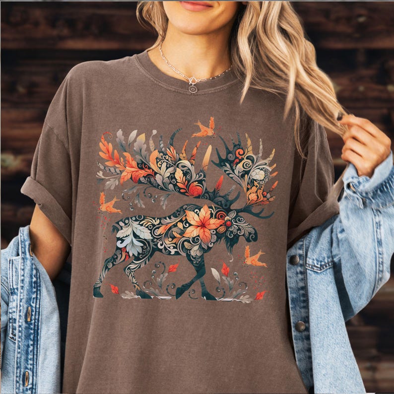 May include: Brown t-shirt featuring a detailed floral moose design with orange, green, and blue accents. The moose has large antlers and is surrounded by stylized leaves and birds. The shirt has a relaxed fit.