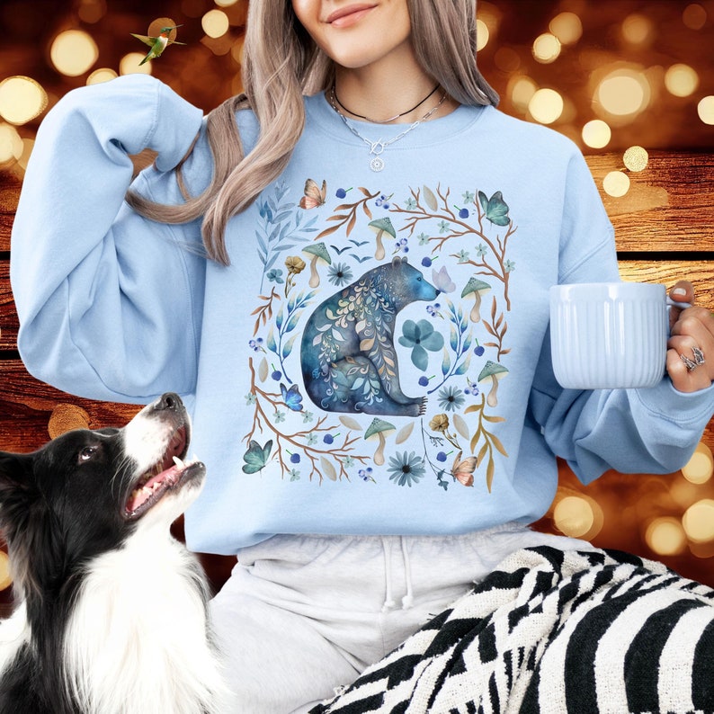 May include: A light blue sweatshirt with a watercolor illustration of a bear sitting in a circle of flowers and leaves. The bear is blue and gray with intricate details. The flowers are in shades of blue, pink, and yellow.