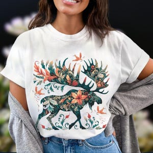 May include: White t-shirt featuring a detailed moose design with floral and leaf patterns in teal, orange, and green. The moose has large antlers and is surrounded by small birds. The design is centered on the shirt.