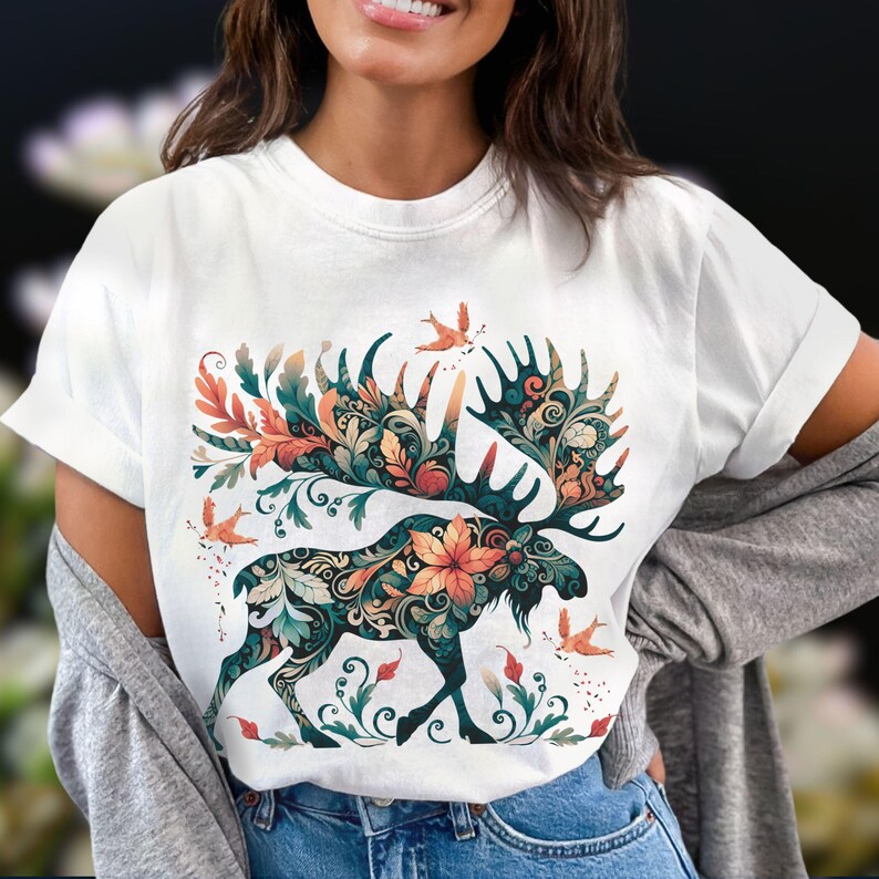 May include: White t-shirt featuring a detailed moose design with floral and leaf patterns in teal, orange, and green. The moose has large antlers and is surrounded by small birds. The design is centered on the shirt.