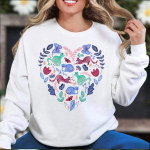 May include: White sweatshirt featuring a heart-shaped design filled with colorful illustrations. The design includes cats, butterflies, and floral elements in shades of blue, pink, green, and purple. The sweatshirt is a casual, comfortable garment.