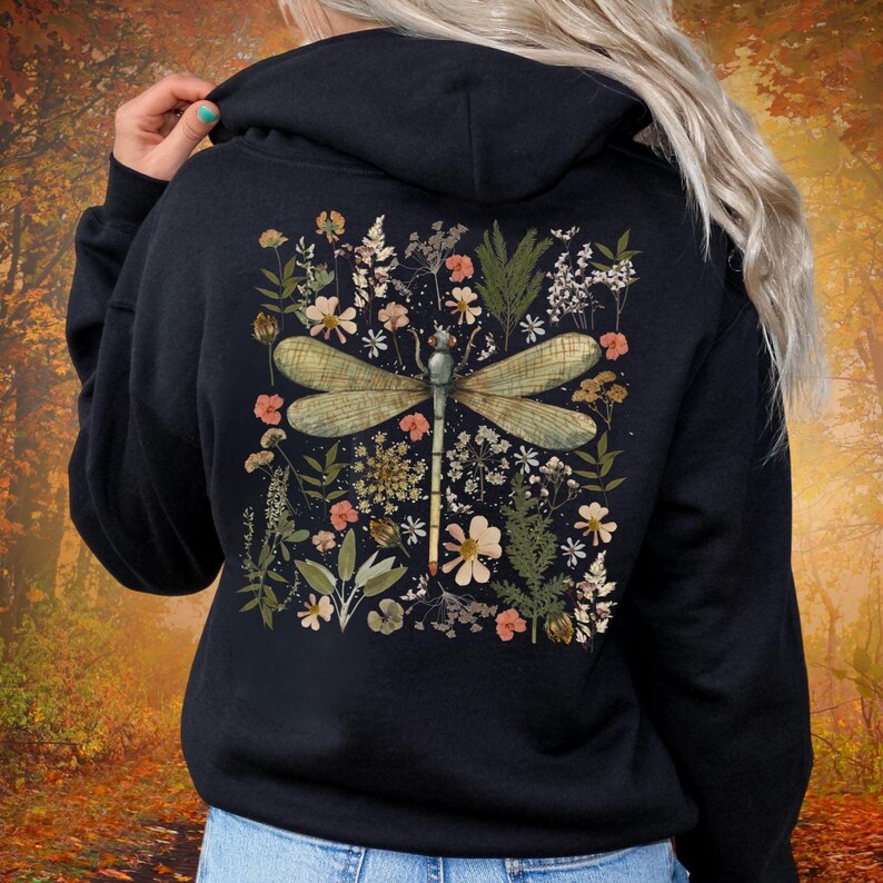 May include: Black hoodie featuring a detailed illustration of a dragonfly surrounded by flowers and foliage. The dragonfly has a green and tan color scheme, and the flowers are in shades of pink, white, and yellow. The design is centered on the back of the hoodie.