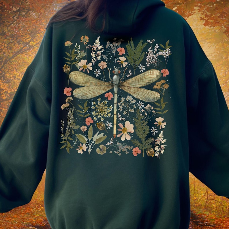 May include: Dark green hoodie featuring a large dragonfly design with floral accents. The dragonfly has detailed wings and is surrounded by various flowers and foliage in shades of pink, white, and green. The hoodie is a solid dark green color.