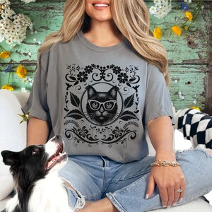 May include: A gray t-shirt with a black cat wearing glasses in a floral frame design.