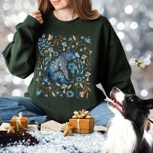 May include: A dark green sweatshirt with a blue bear surrounded by flowers and mushrooms. The bear is sitting on its haunches and has a floral pattern on its body. The sweatshirt is a comfortable and stylish choice for anyone who loves nature.