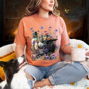 May include: A woman wearing a peach colored t-shirt with a raven, potion bottles, and flowers printed on the front. The raven is wearing a witch's hat.