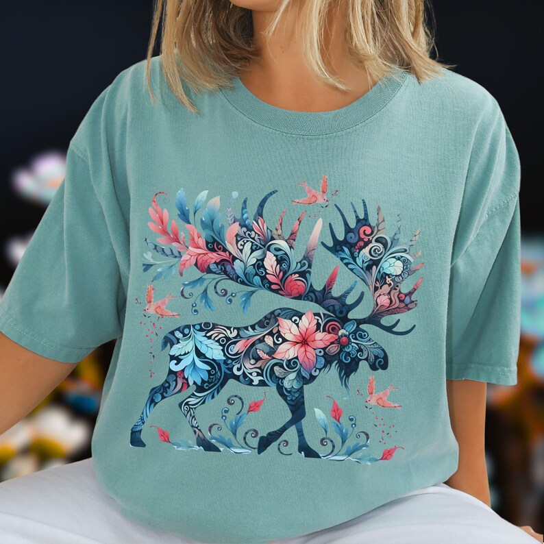 May include: A teal-colored t-shirt featuring a detailed illustration of a moose. The moose is adorned with a floral design in shades of pink, blue, and white. The antlers are dark blue, and small birds are scattered around the moose.