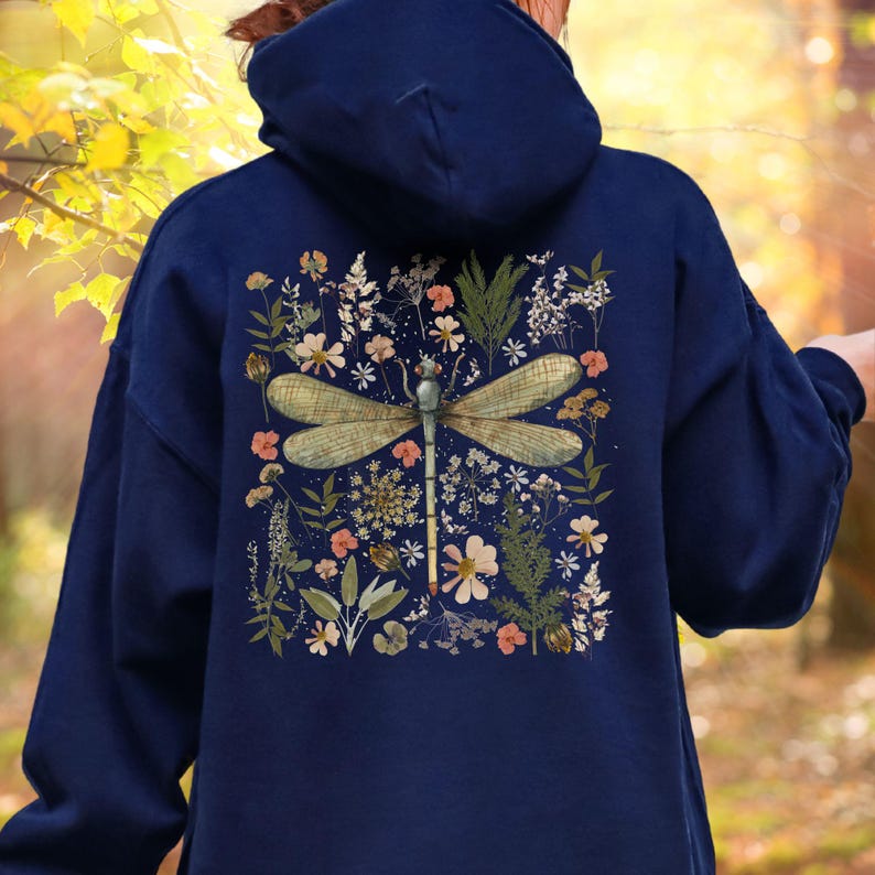 May include: Navy blue hoodie featuring a detailed illustration of a dragonfly with delicate wings, surrounded by a floral arrangement of wildflowers in shades of pink, white, and green. The design is centered on the back of the hooded sweatshirt.
