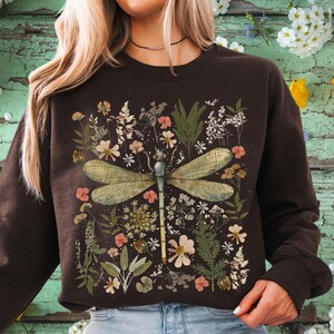 May include: A dark brown sweatshirt featuring a large dragonfly design with detailed wings, surrounded by a collage of flowers and foliage in shades of pink, white, and green. The design is centered on the front of the sweatshirt.