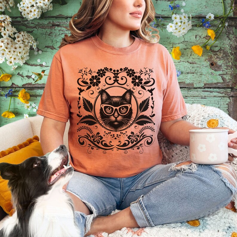 May include: A woman wearing a peach-colored t-shirt with a black cat wearing glasses graphic. The graphic is surrounded by black floral designs. The woman is sitting on a couch with a dog beside her.