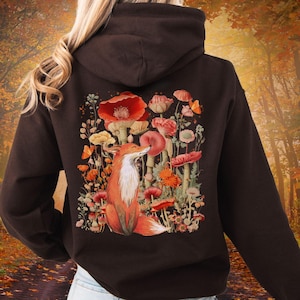 May include: A dark brown hoodie featuring a vibrant illustration of a fox amidst colorful mushrooms and flowers. The design includes red, orange, and yellow hues, creating a whimsical, nature-inspired aesthetic. The hoodie has a hood and long sleeves.