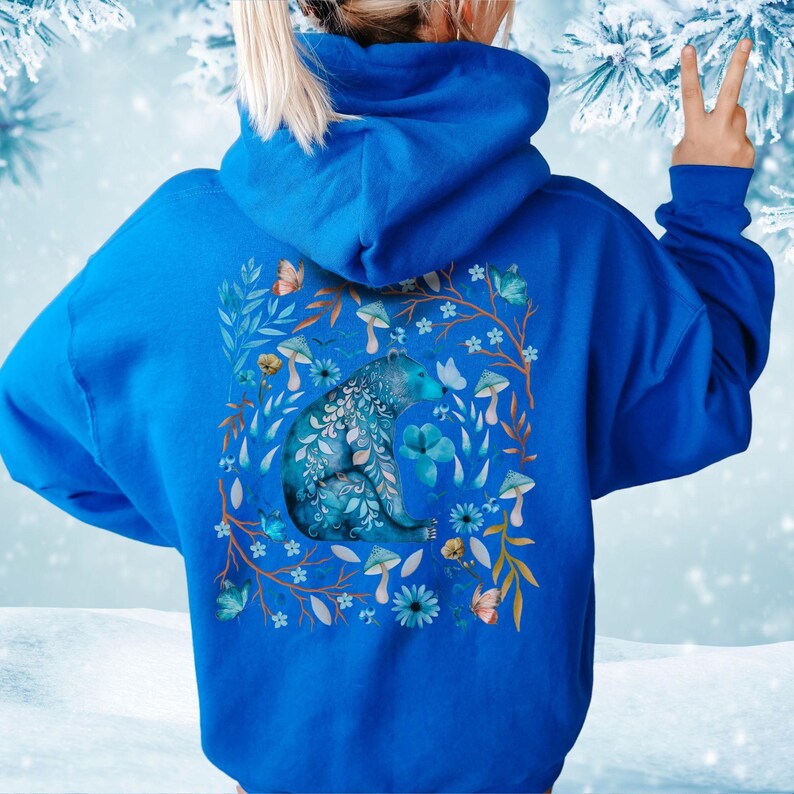 May include: A royal blue hoodie featuring a detailed illustration of a bear with floral and mushroom accents. The bear is surrounded by flowers, butterflies, and mushrooms in shades of blue, orange, and white. The hoodie has a drawstring hood.