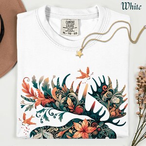 May include: White t-shirt with a colorful floral and antler design. The design incorporates orange, teal, and green leaves and flowers. A gold star pendant necklace rests on the shirt. The shirt is labeled XL-TG-EG.