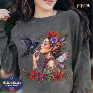May include: A charcoal gray long-sleeve sweatshirt featuring a graphic print of a fairy with pointed ears and wings, interacting with a black crow. The design includes colorful red, purple, and orange flowers, brown acorns, and thin branches.