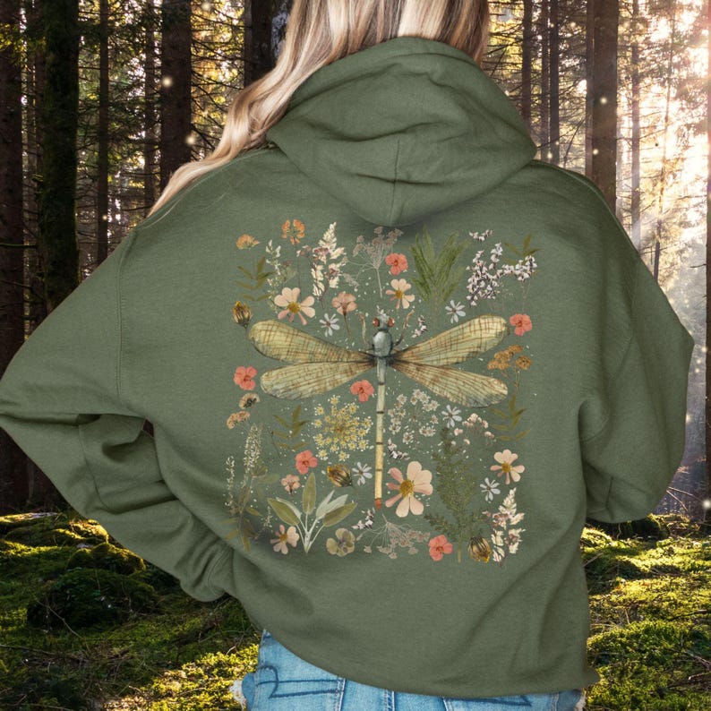 May include: Olive green hoodie featuring a large dragonfly design on the back, surrounded by floral and botanical elements in shades of peach, white, and green. The hoodie has a drawstring hood and a kangaroo pocket.