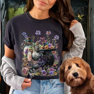 May include: A black t-shirt with a graphic print of a raven wearing a witch's hat surrounded by flowers, bottles, and other magical items.