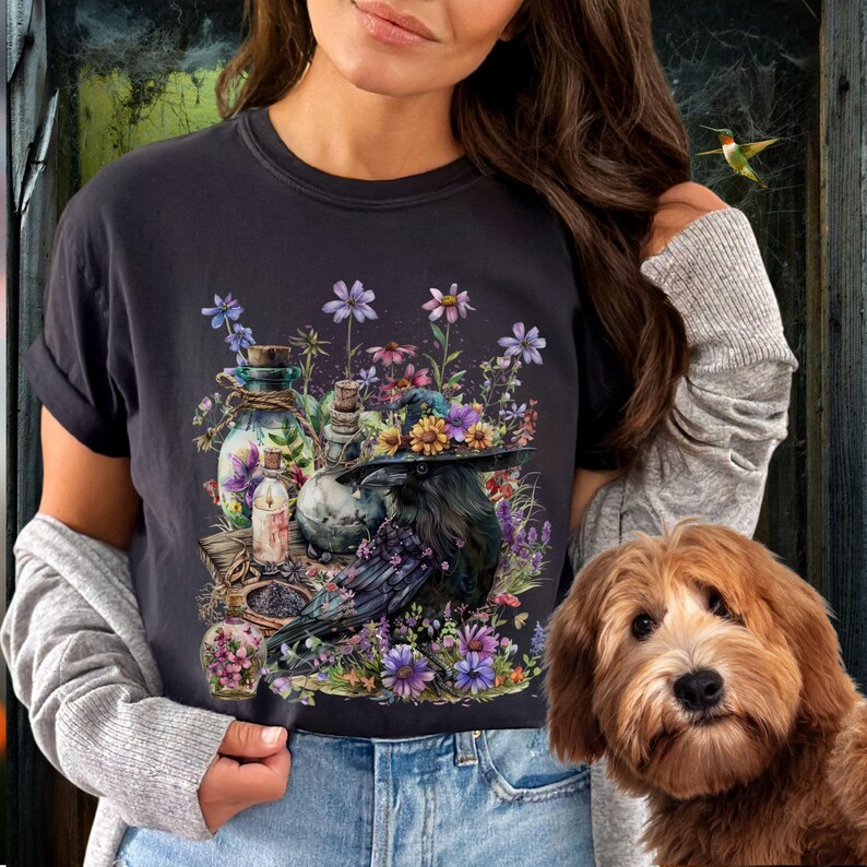 May include: A black t-shirt with a graphic print of a raven wearing a witch's hat surrounded by flowers, bottles, and other magical items.