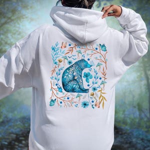 May include: White hoodie featuring a watercolor design of a blue bear surrounded by flowers, butterflies, and mushrooms. The bear is the focal point, with intricate floral details. The design is on the back of the hoodie.