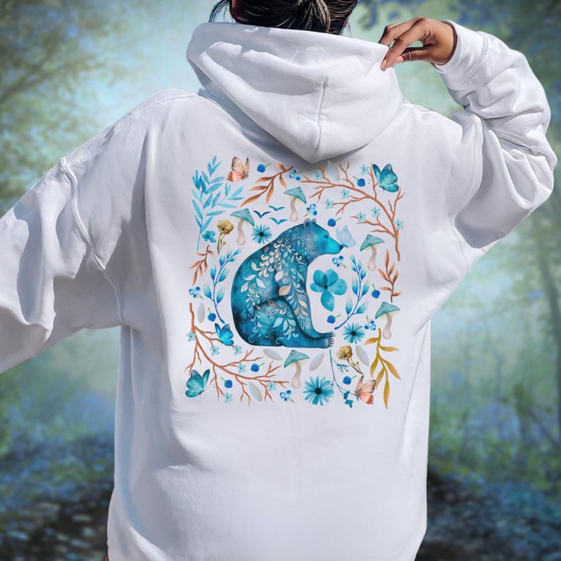 May include: White hoodie featuring a watercolor design of a blue bear surrounded by flowers, butterflies, and mushrooms. The bear is the focal point, with intricate floral details. The design is on the back of the hoodie.