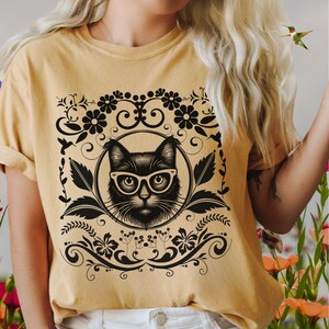 May include: A yellow t-shirt with a black cat wearing glasses and a floral design. The cat is surrounded by a circular frame with swirls and leaves.