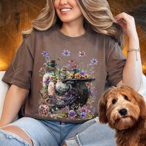 May include: A brown t-shirt with a graphic of a raven wearing a witch's hat surrounded by flowers and potion bottles.