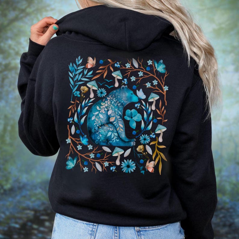 May include: Black hoodie featuring a blue bear design surrounded by flowers, mushrooms, and butterflies. The bear is intricately detailed with floral patterns. The design is set against a backdrop of leaves and branches.