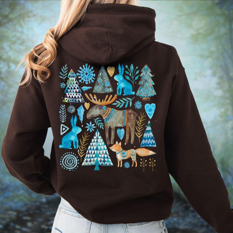 May include: A dark brown hoodie featuring a whimsical design on the back. The design includes blue rabbits, Christmas trees, a moose, and a fox, with accents of gold and white. The hoodie has a hood and a relaxed fit.