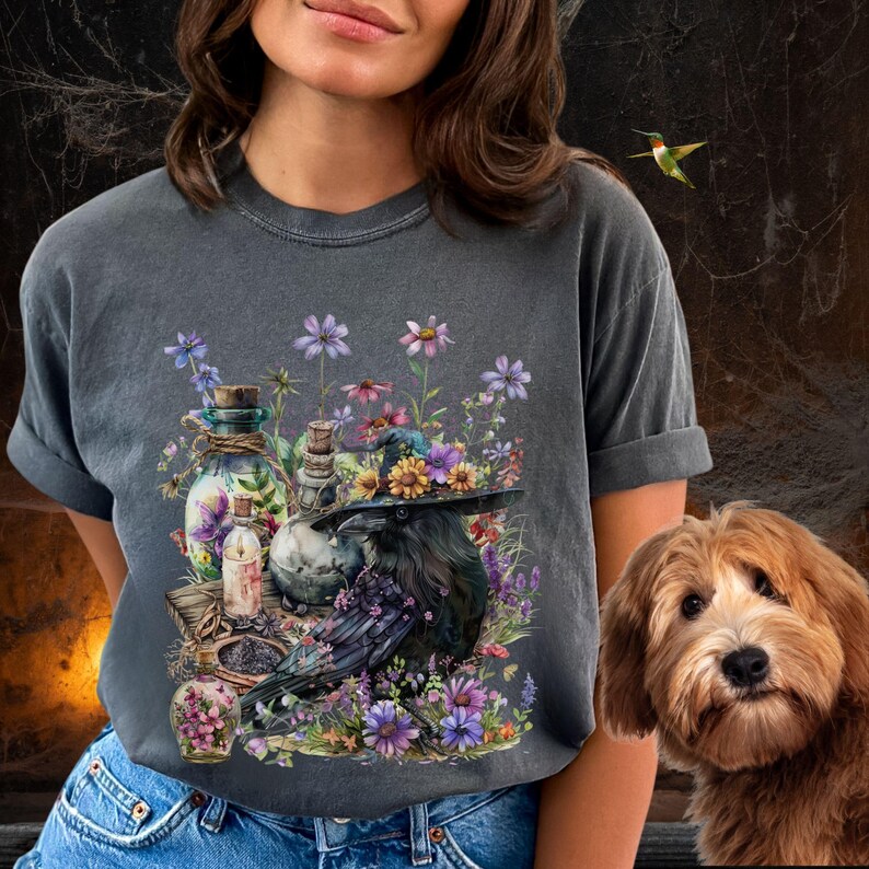 May include: A dark gray t-shirt with a black raven wearing a witch's hat adorned with flowers. The raven is surrounded by various bottles, jars, and candles, all set against a backdrop of purple and pink flowers.