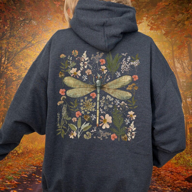May include: Dark navy blue hoodie featuring a large dragonfly design on the back. The dragonfly is surrounded by a floral arrangement with peach and white flowers, green leaves, and other botanical elements. The hoodie has a drawstring hood.