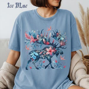 May include: Ice blue t-shirt featuring a detailed illustration of a moose with floral antlers and decorative patterns. The design incorporates pink, blue, and green elements, with the text "Ice Blue" at the top left. The shirt is a comfortable, casual style.