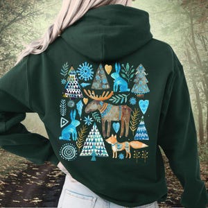May include: Dark green hoodie featuring a whimsical forest scene. The design includes blue rabbits, trees, a moose, and a fox, all in a folk art style. The artwork is centered on the back of the hoodie, creating a cozy, nature-inspired aesthetic.