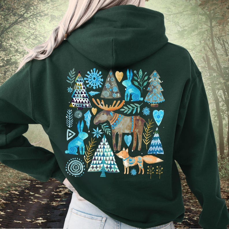 May include: Dark green hoodie featuring a whimsical forest scene. The design includes blue rabbits, trees, a moose, and a fox, all in a folk art style. The artwork is centered on the back of the hoodie, creating a cozy, nature-inspired aesthetic.