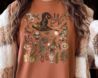 May include: Rust-colored sweatshirt featuring a detailed illustration of a witch's hat, flowers, mushrooms, and other botanical elements. The design includes a skull, butterflies, and a candle. The overall theme is a whimsical, nature-inspired design.