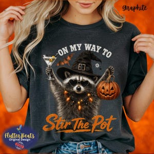May include: A charcoal gray t-shirt featuring a graphic of a raccoon wearing a black witch hat and heart-shaped sunglasses. The raccoon holds a martini glass and a jack-o'-lantern. Text reads ON MY WAY TO Stir The Pot with a bat above.