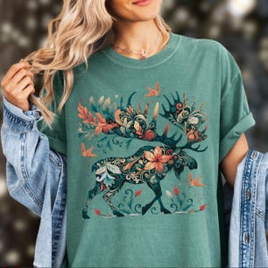May include: A teal-colored t-shirt featuring a detailed illustration of a moose adorned with floral and leaf patterns in shades of orange, teal, and cream. The moose is surrounded by stylized birds and foliage, creating a nature-inspired design.