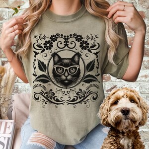 May include: A green t-shirt with a black cat wearing glasses and a floral design. The cat is surrounded by a decorative frame.